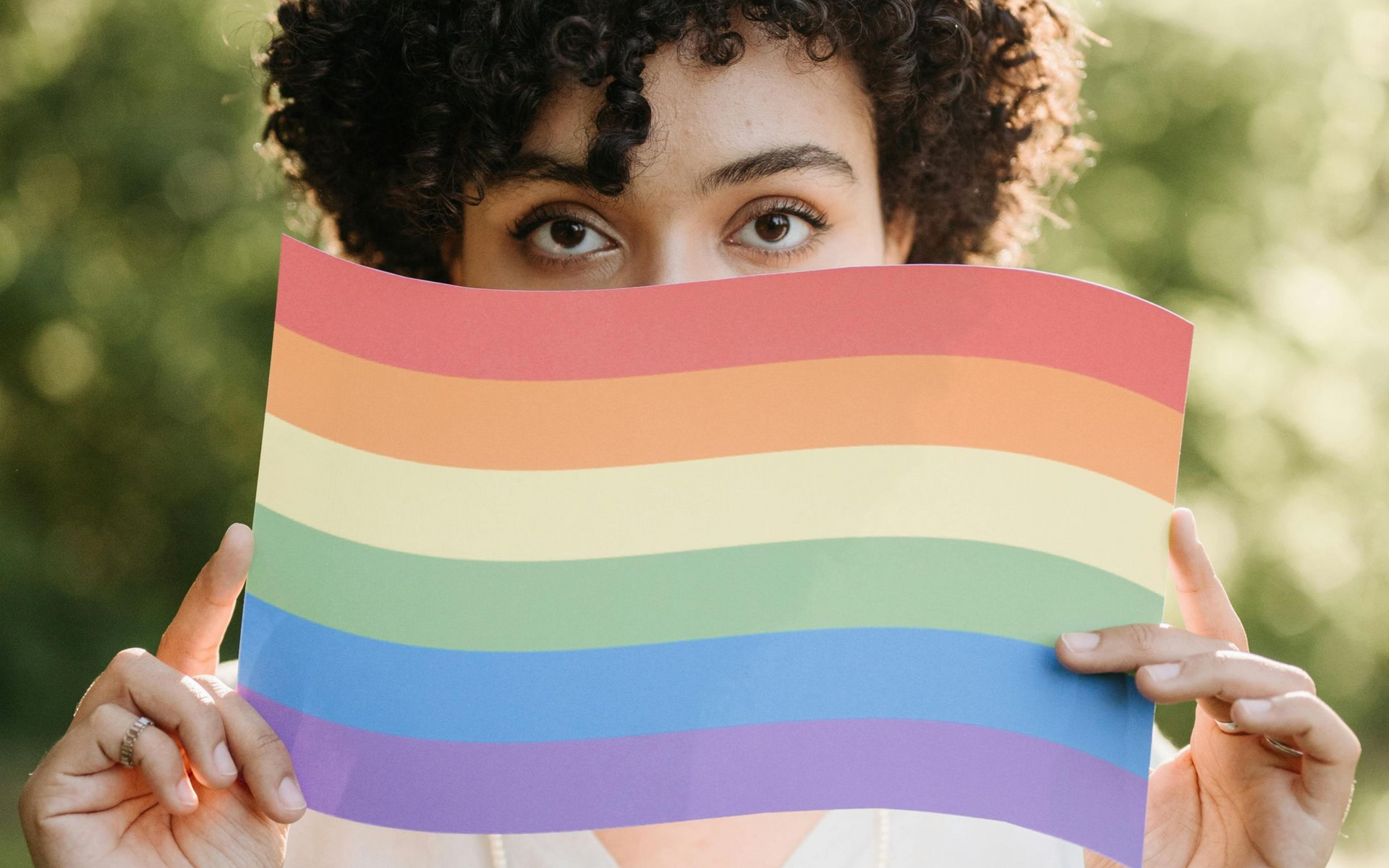 A person holds a rainbow flag in front of their face, with their eyes visible above the flag.