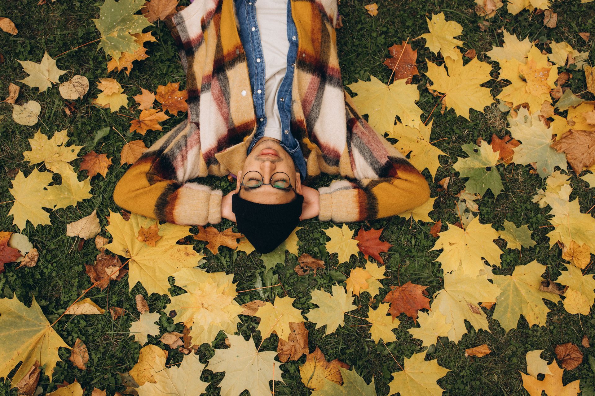 A person lays on their back in on grass, surrounded by scattered yellow and orange leaves.