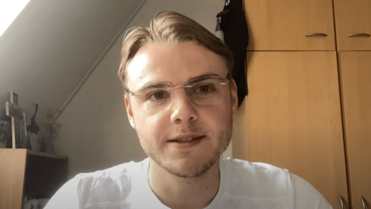 Een persoon met een bril zit binnen voor houten kasten en een raam en kijkt naar de camera.