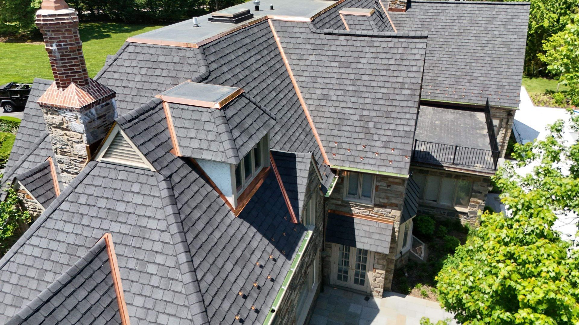Aerial view of a house with gray shingled roofs, a chimney, and a leafy green yard.