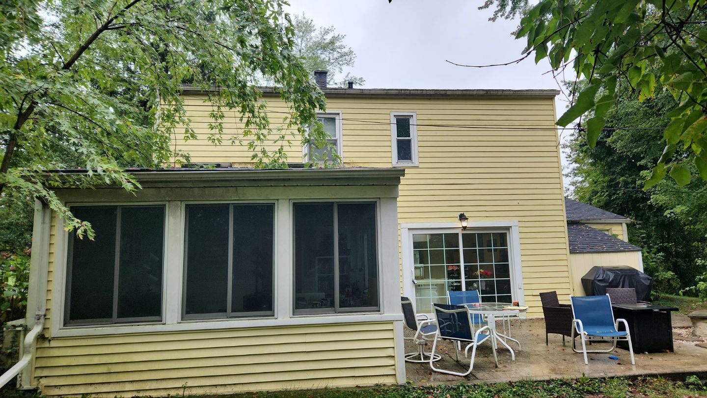 A yellow two-story house with a screened-in porch, a patio with chairs, and lush green trees in the background.