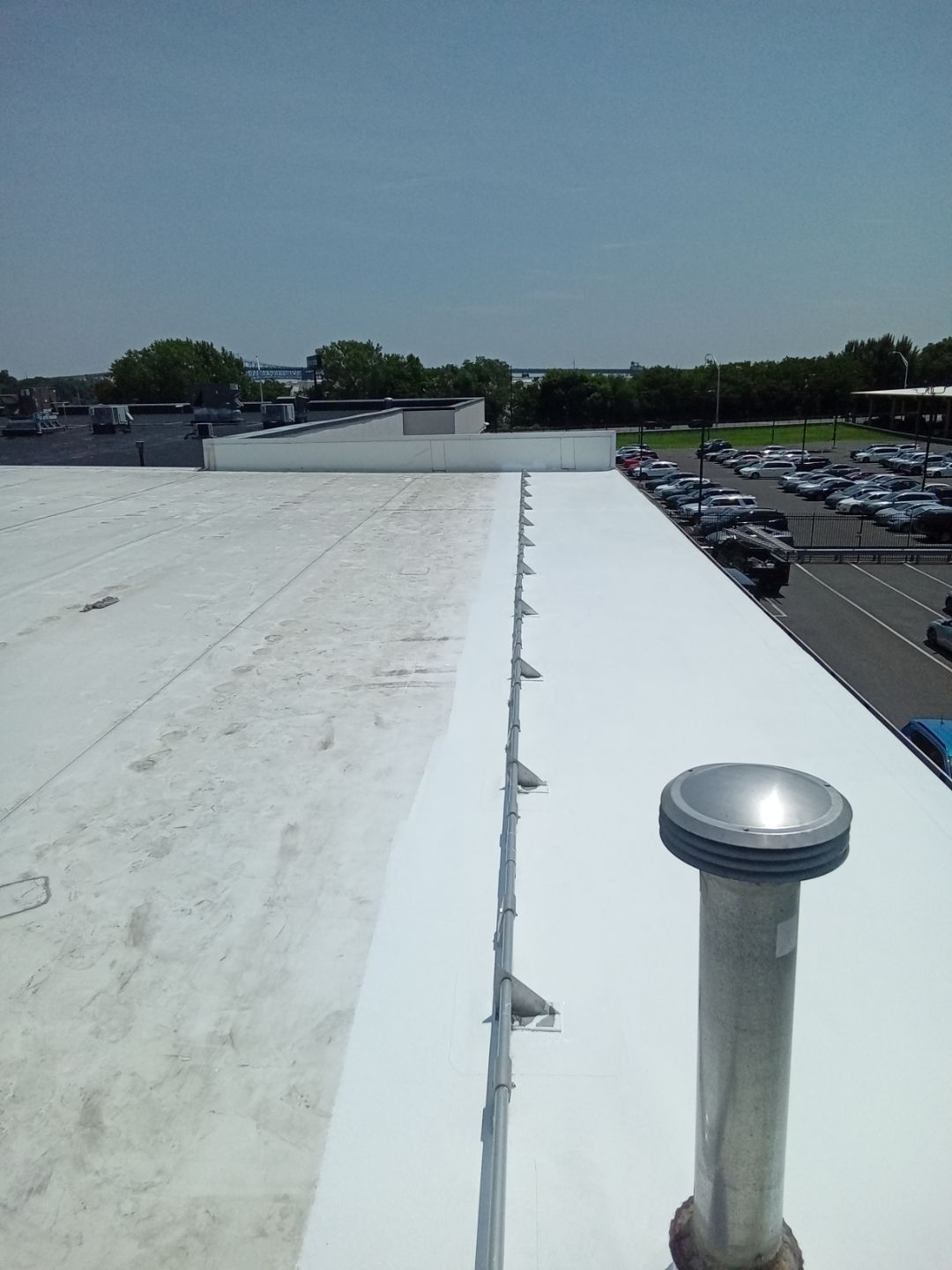 Rooftop with white membrane roof, vent pipe, and parking lot in the distance under a clear sky