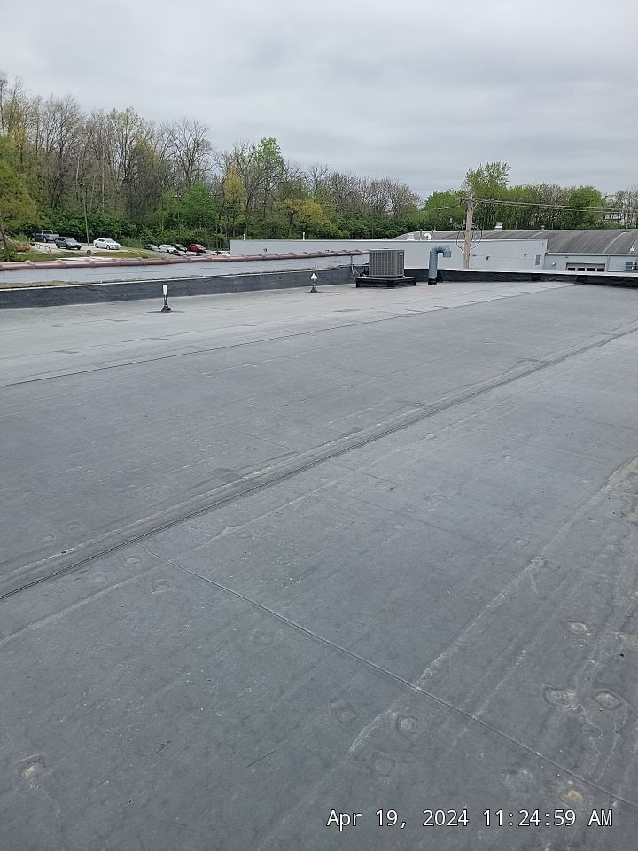 Flat rooftop with gray membrane, HVAC units, and trees beyond a low parapet under cloudy sky
