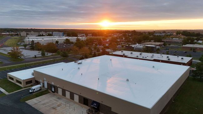 Aerial view of a large white-roofed building at sunset, with surrounding parking lots and trees.