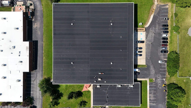 Aerial view of a large building with a dark roof, parking lot, and surrounding trees.
