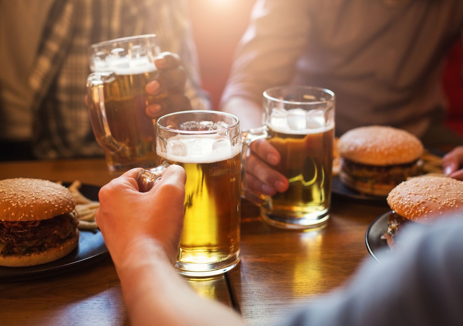 People at a table clink beer glasses, burgers nearby. Sunlight.