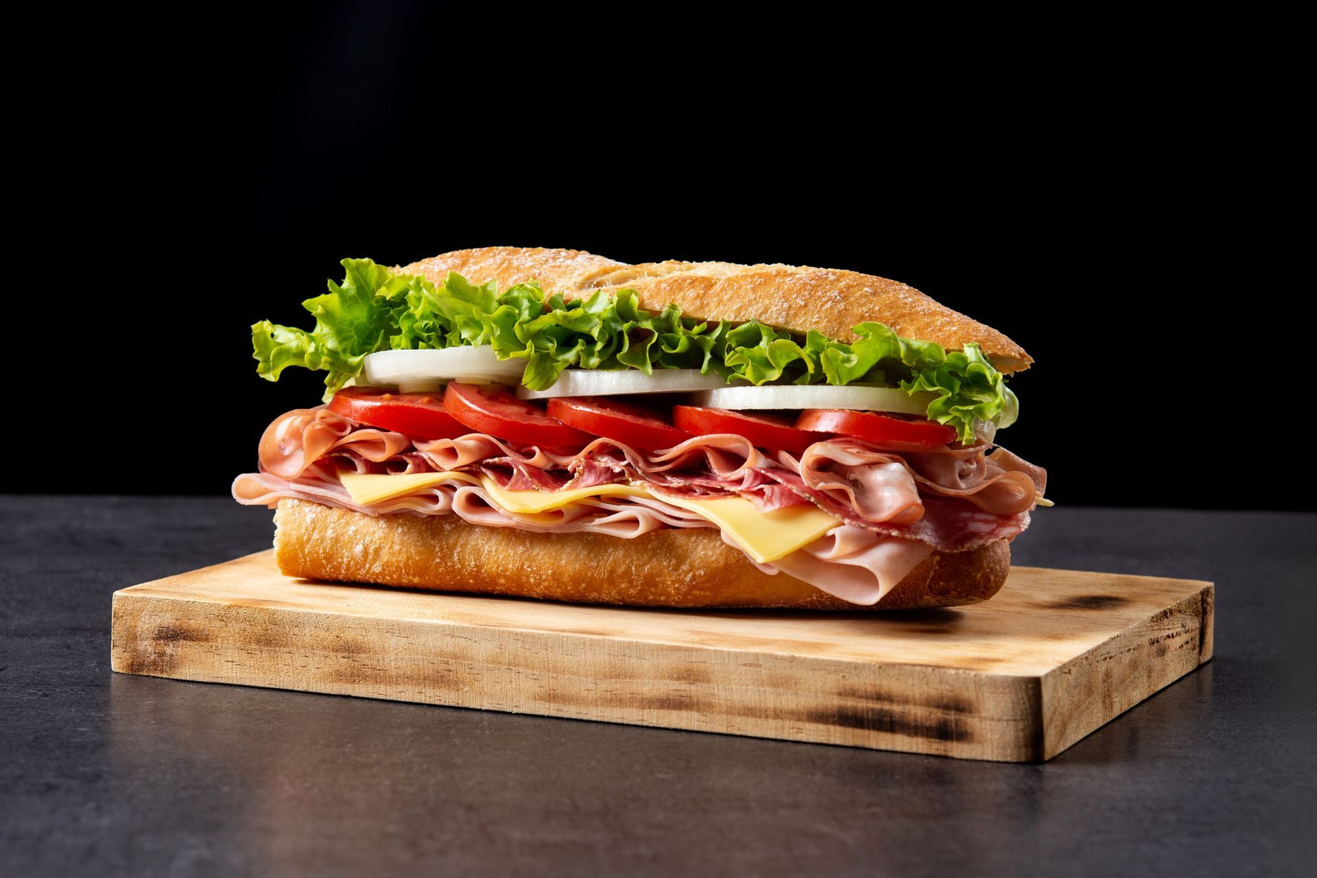 Layered deli sandwich with lettuce, tomato, cheese, and meats on a wooden board against a black background