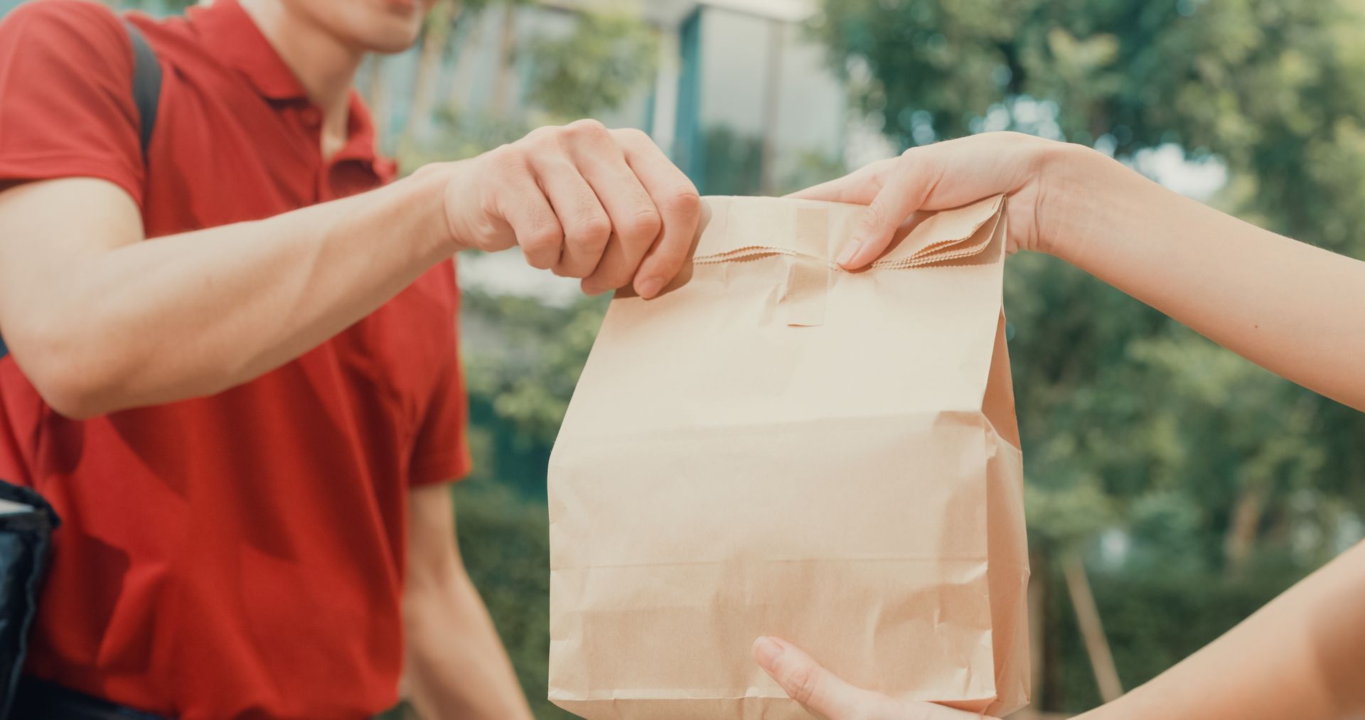 Two people hold a brown paper bag outdoors near a building.