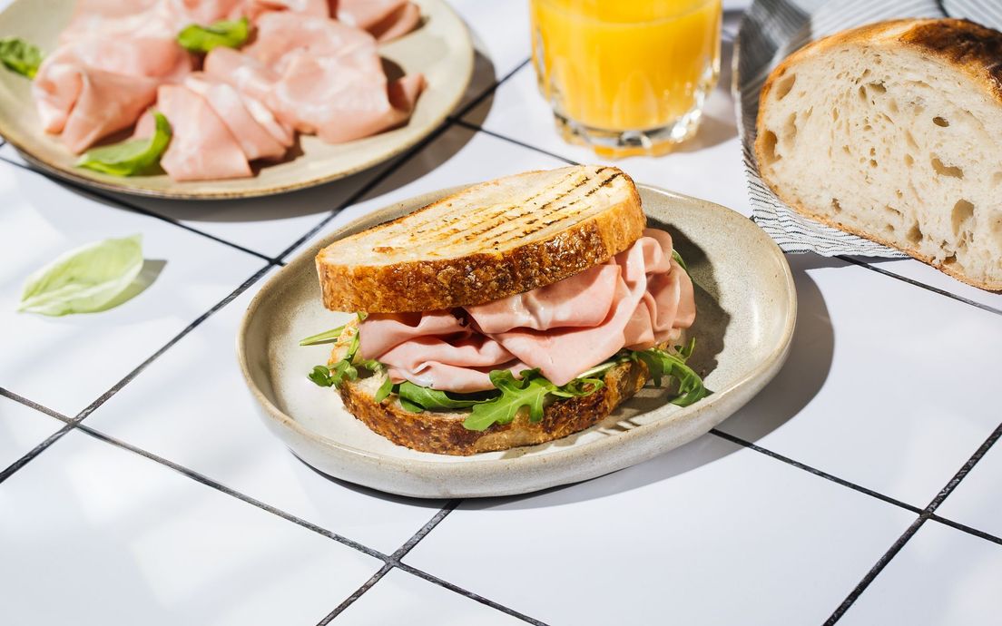 Sandwich on a white plate with sliced meat and greens, beside orange juice and bread on a tiled table
