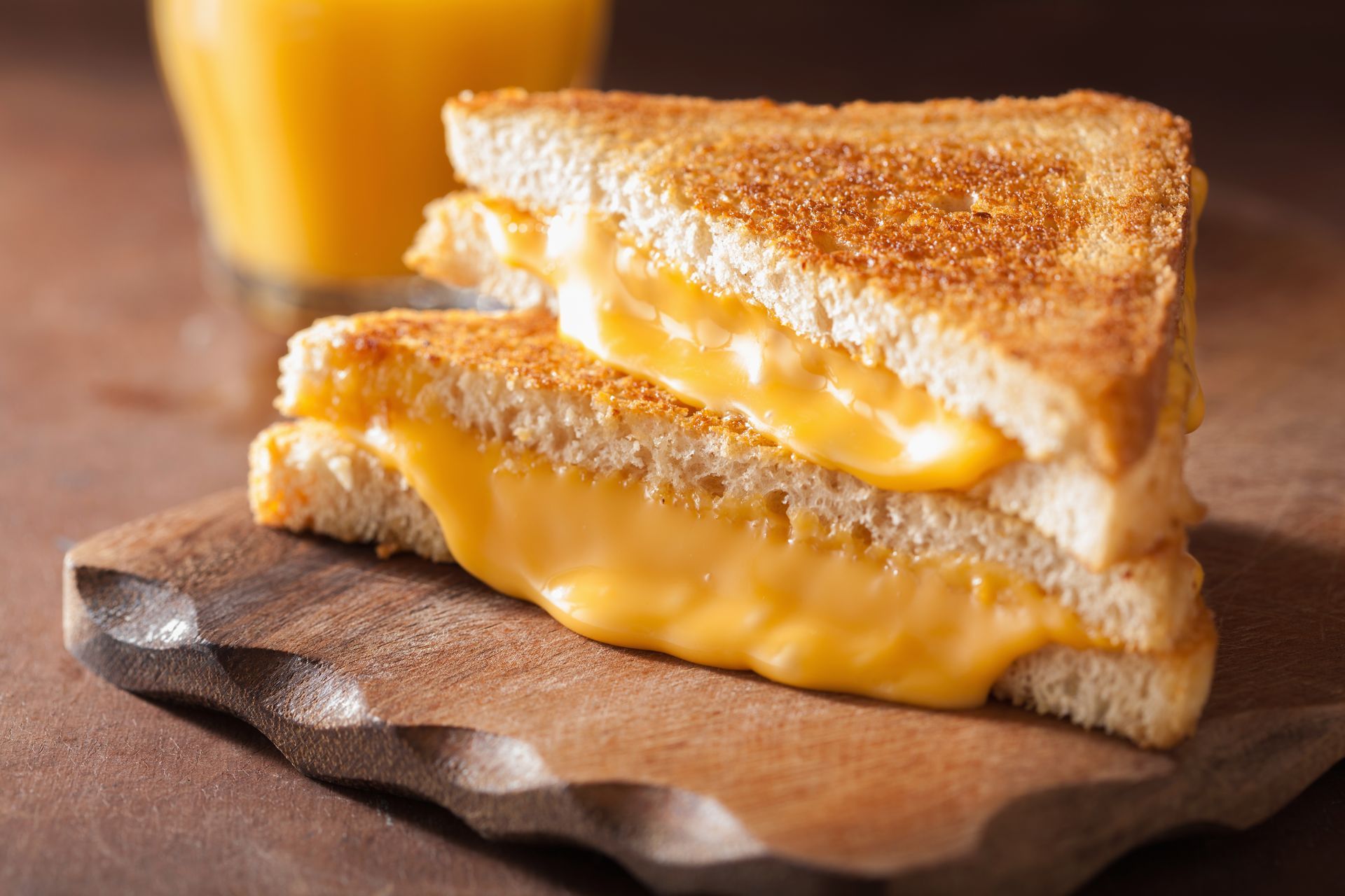 Grilled cheese sandwich with melted cheddar on a wooden board, with a glass of orange juice in the background