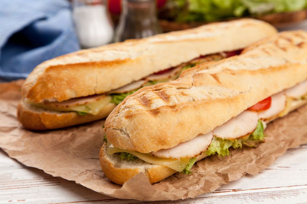 Two baguette sandwiches with lettuce and meat on brown paper on a rustic table