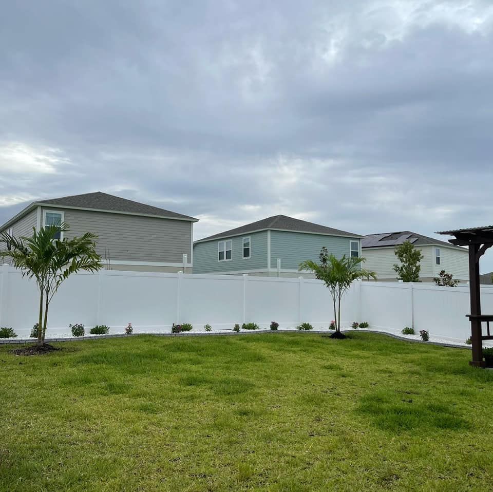 A white fence surrounds a lush green field with houses in the background.