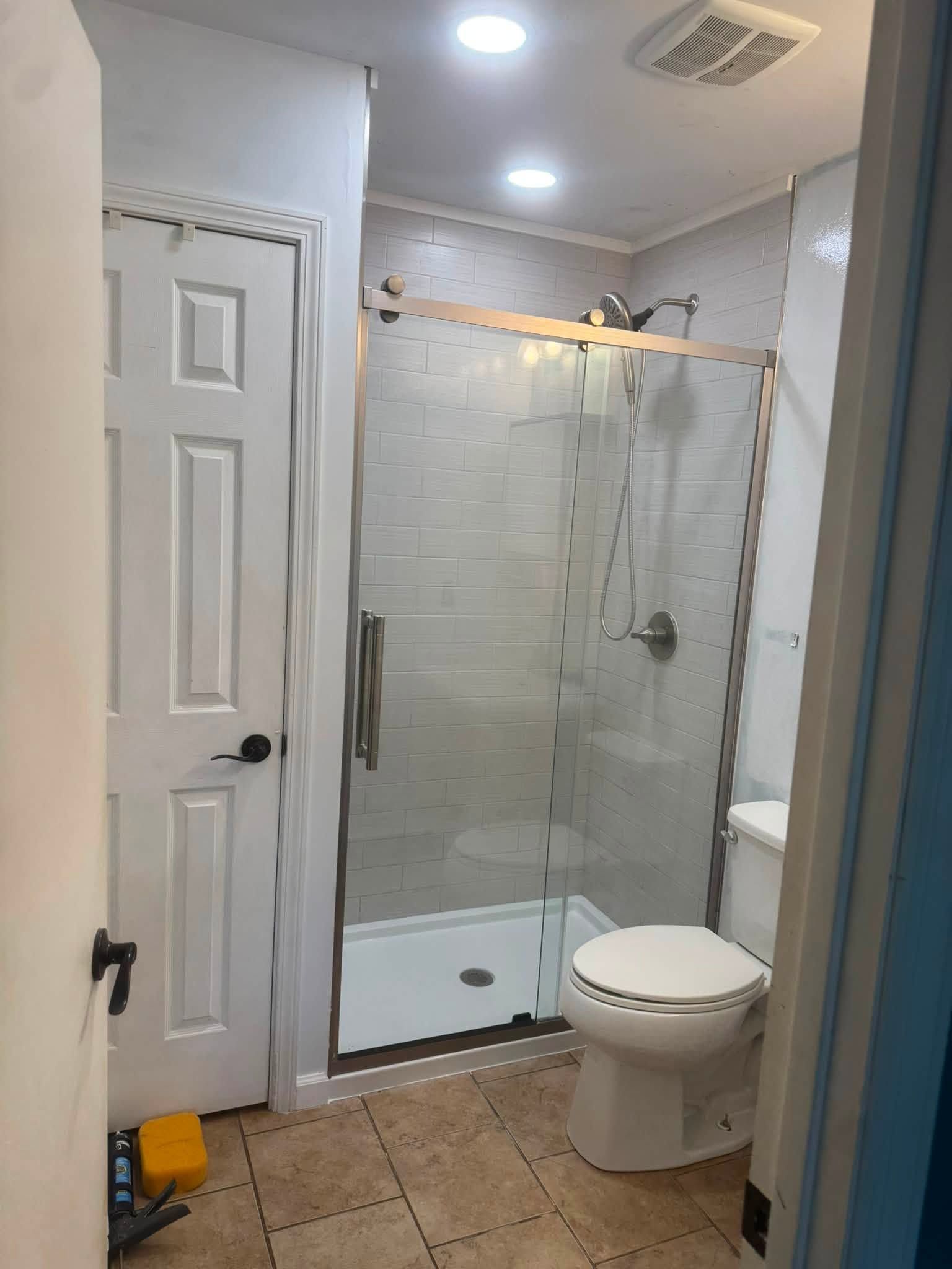 Small bathroom with glass shower, toilet, white doors, and beige tile floor.