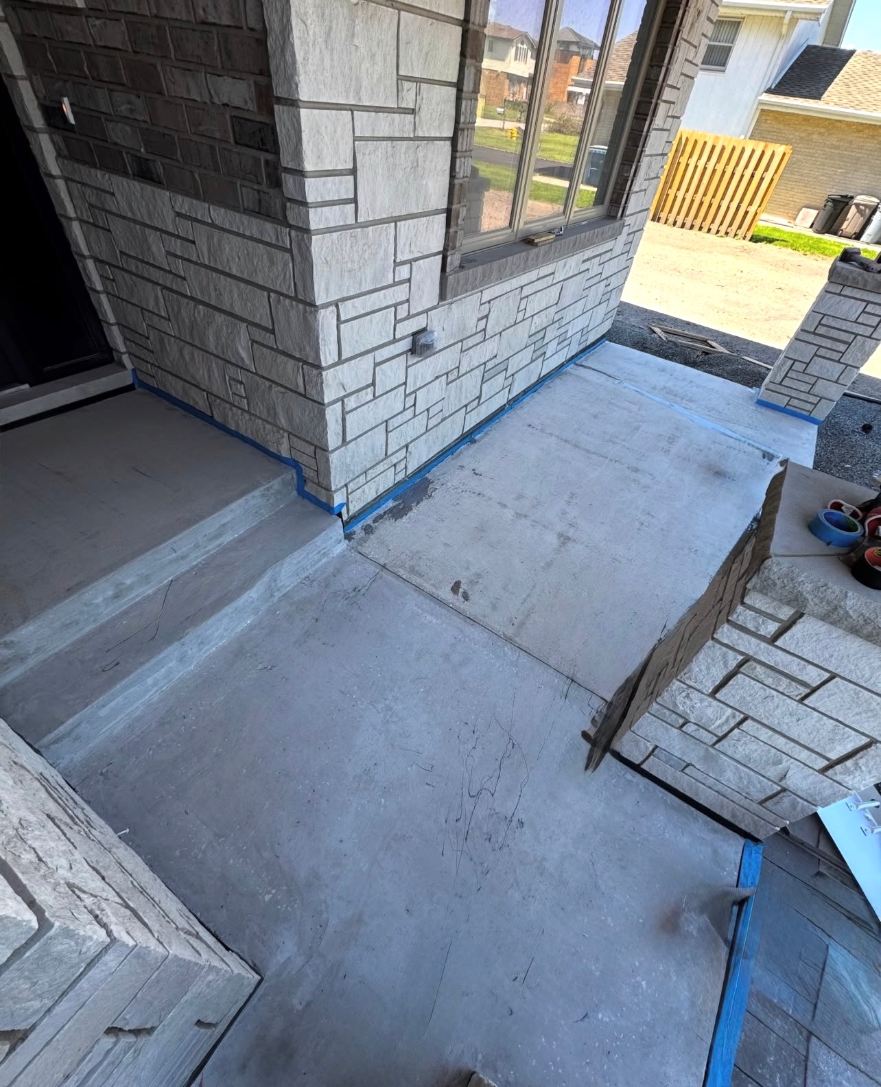 Concrete patio under construction beside a brick house, with steps, a window, and stacked pavers.