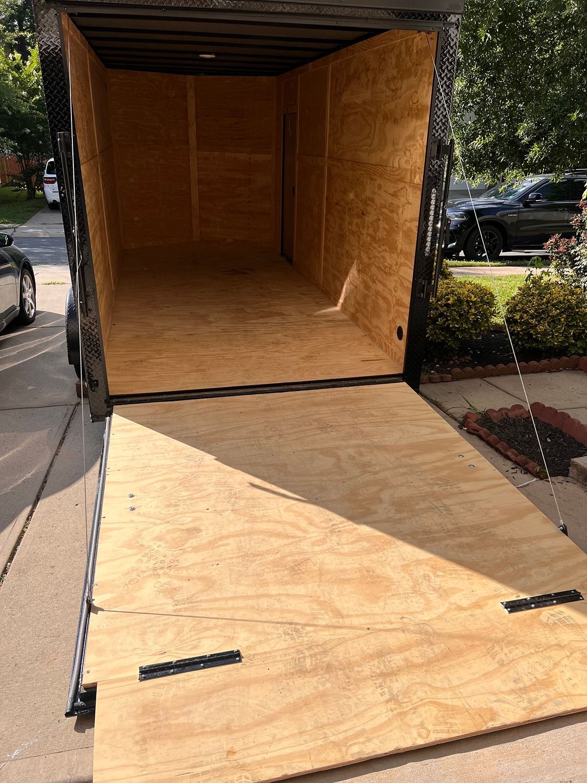 Open empty wooden cargo trailer with ramp lowered onto a driveway, parked beside cars and hedges.