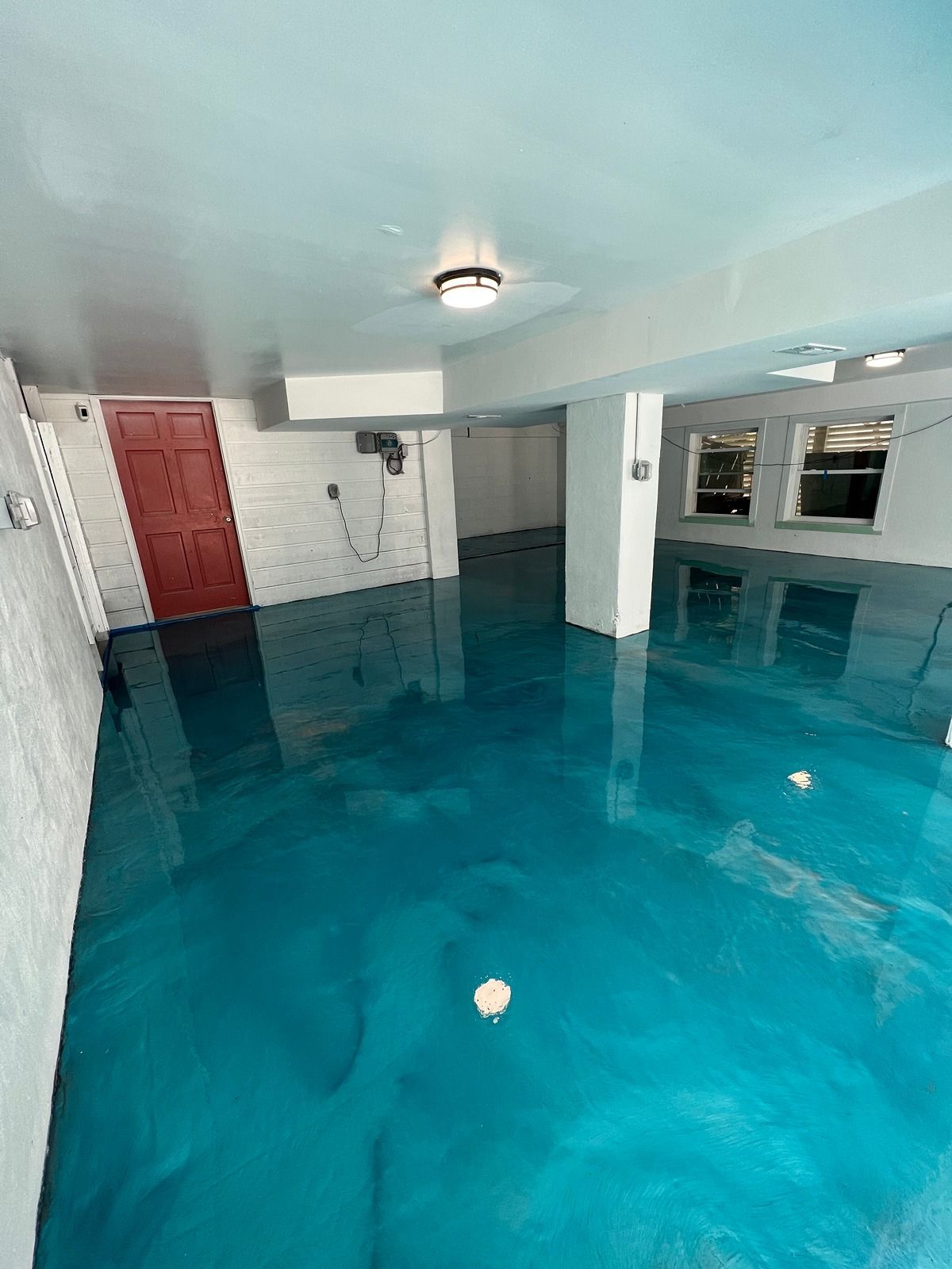 Indoor room flooded with blue-tinted water, with white walls, pillars, and a red door.