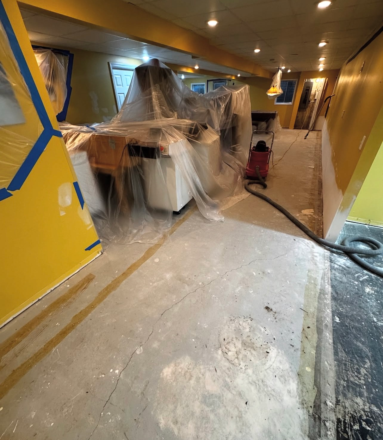 Flooded basement with plastic-covered furniture, debris on the floor, and a hose across the room