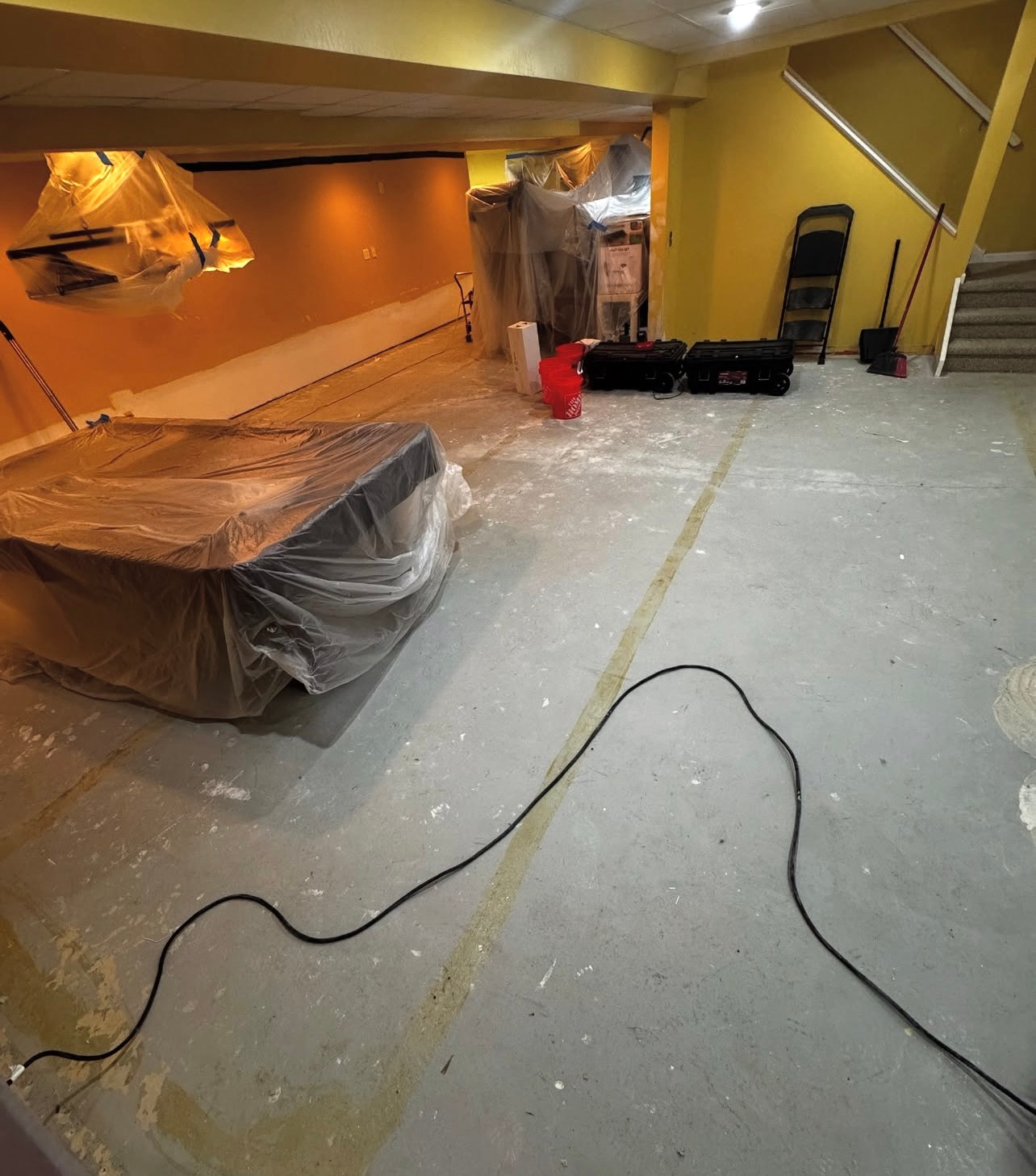 Orange-lit basement with covered furniture, tools, and plastic sheeting over the floor during renovation