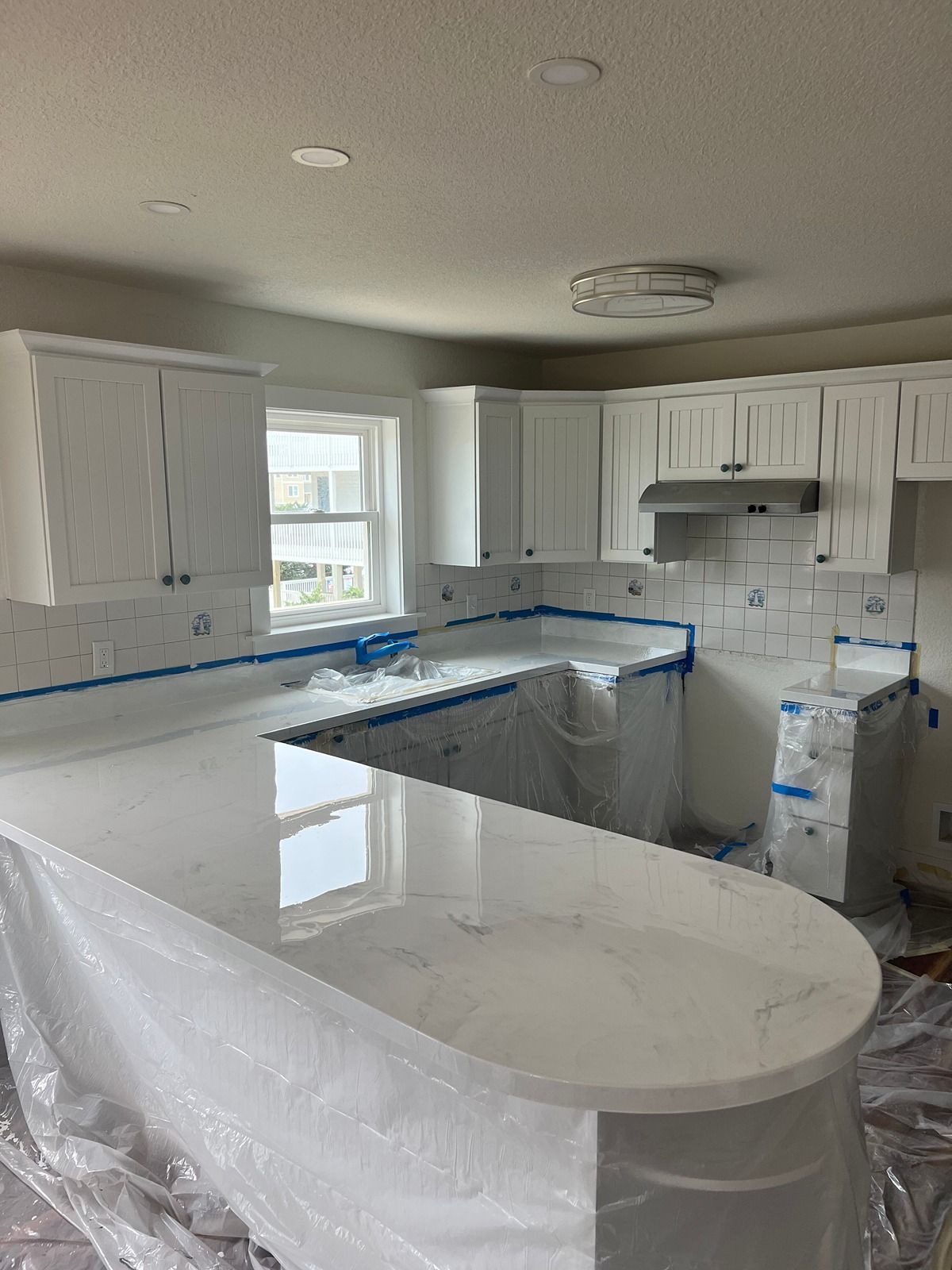 Bright white kitchen with wrapped countertops and cabinets under renovation, with blue painter’s tape visible.