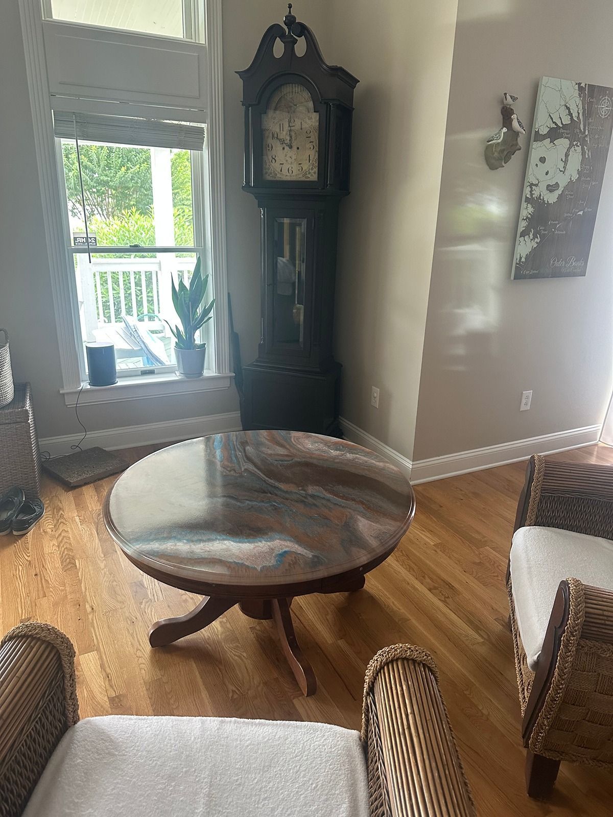 Living room with round wooden table, grandfather clock, wicker chairs, and bright window door.