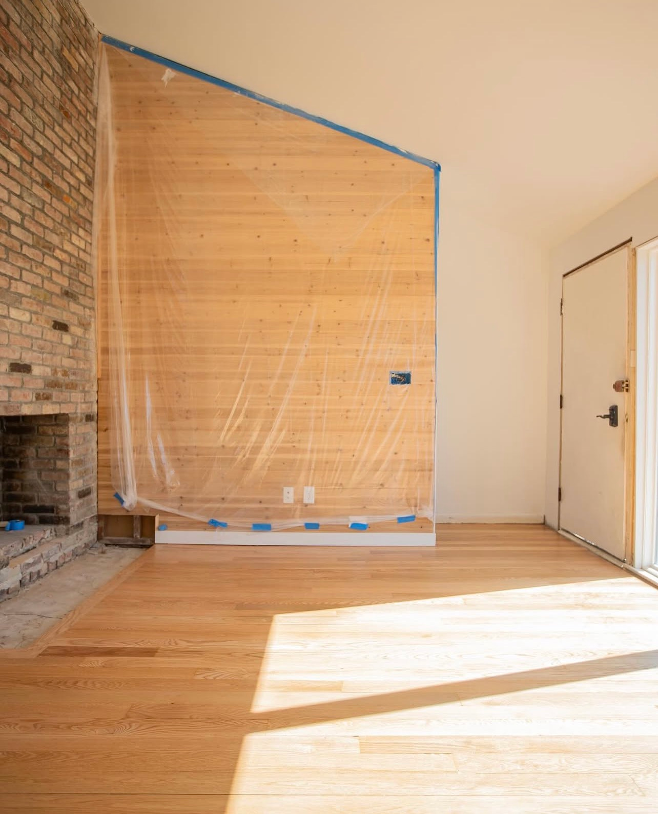 Empty room with hardwood floor, brick fireplace, and a large plywood-covered wall under renovation