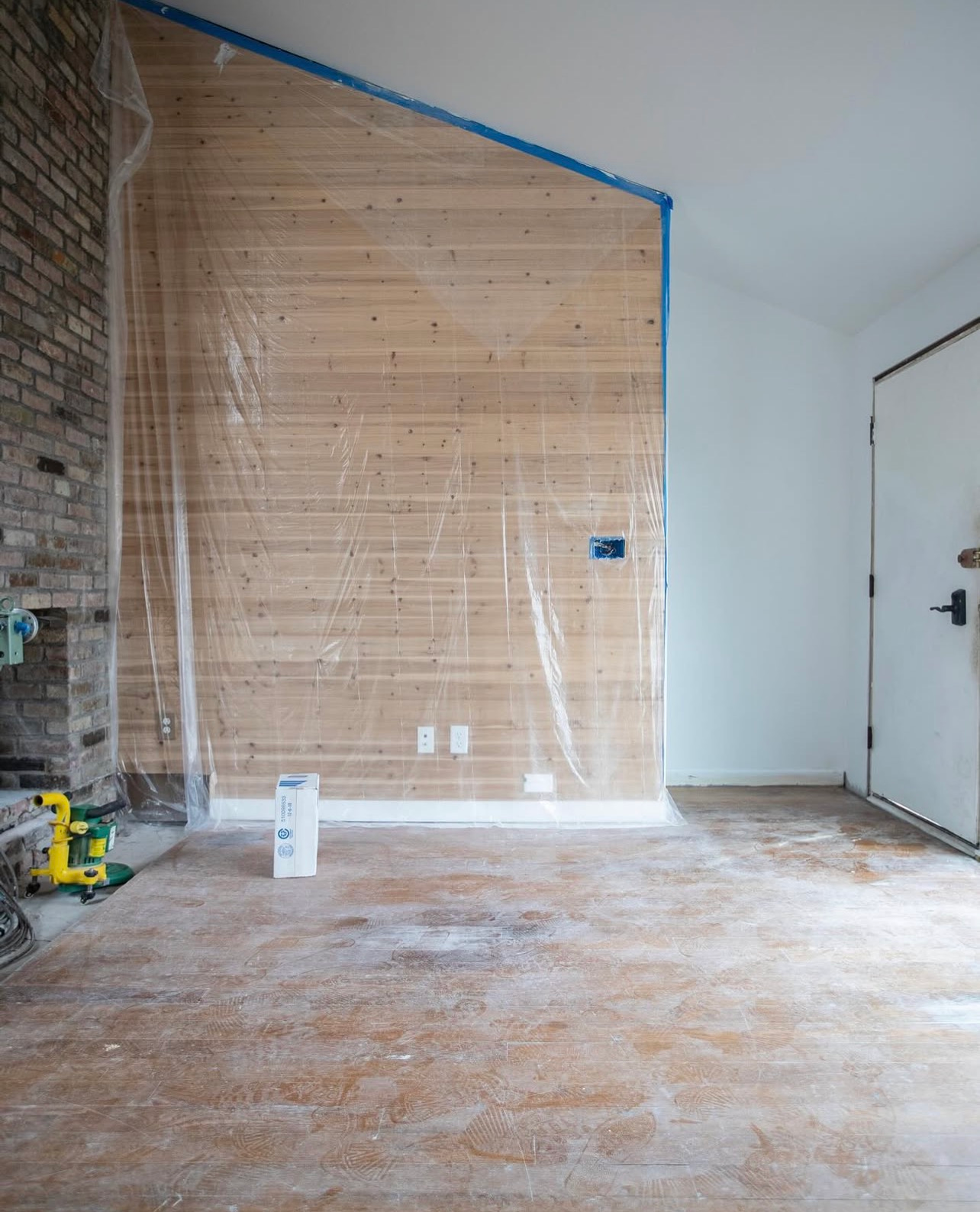 Unfinished room with exposed wall, blue tape, white door, and tools on a dusty floor.