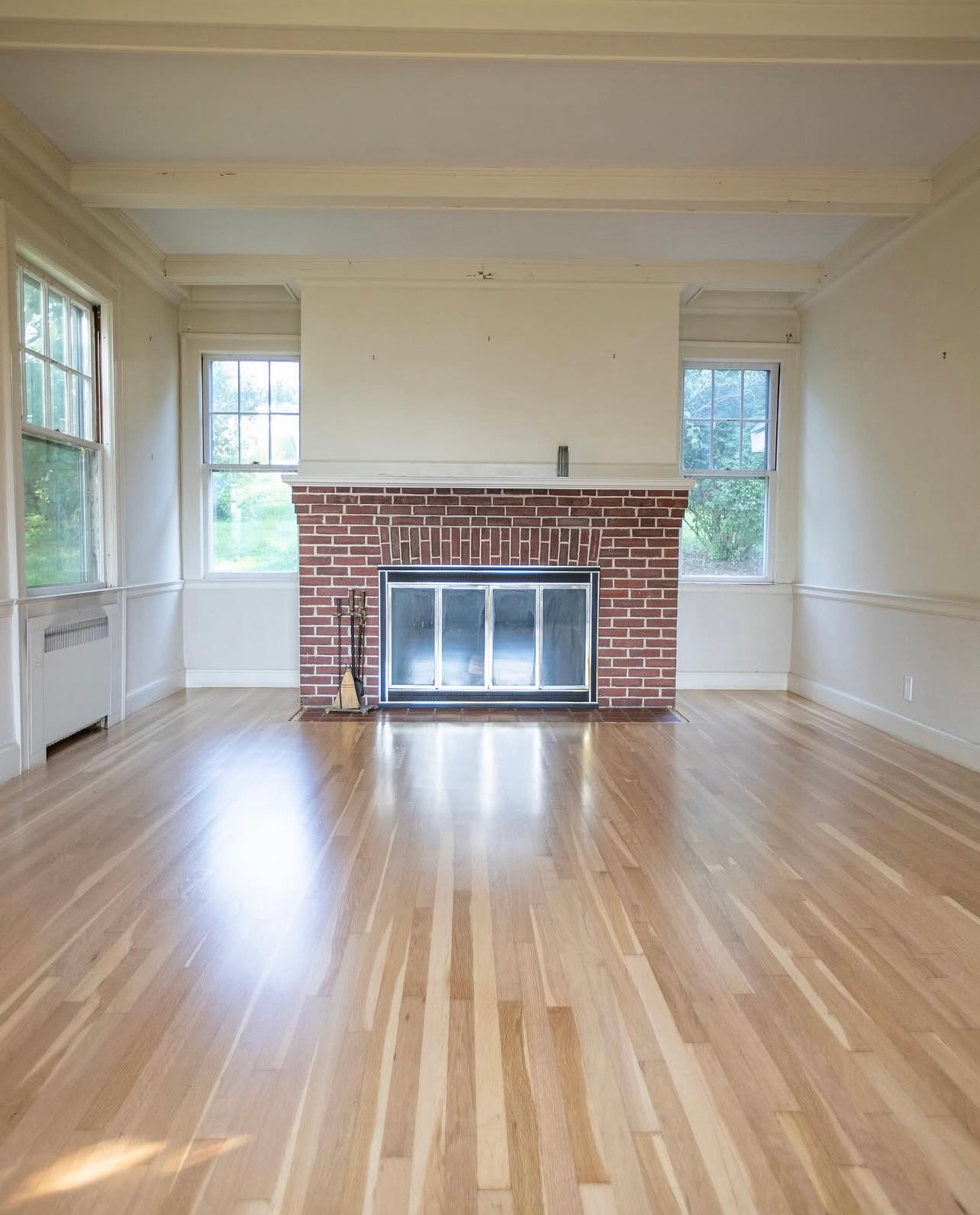 Empty living room with hardwood floors, white walls, and a brick fireplace flanked by windows.