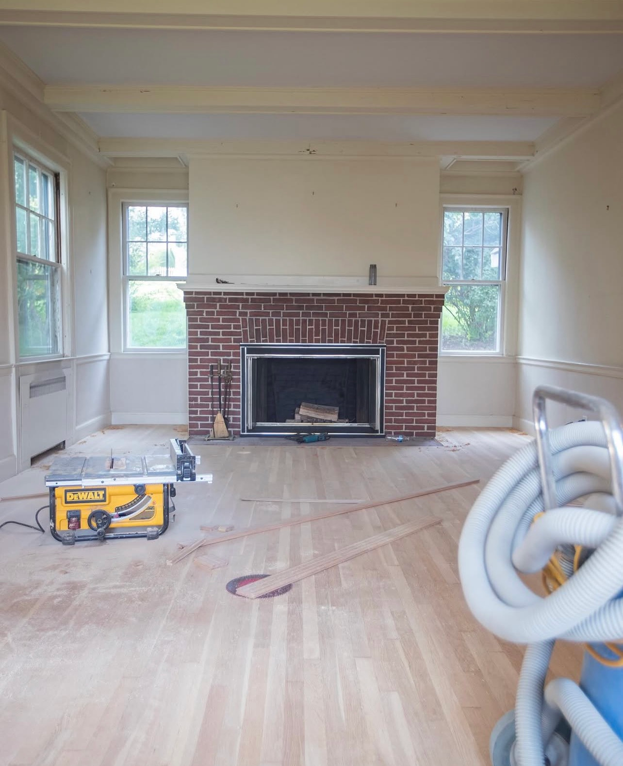 Empty living room with brick fireplace, bare hardwood floor, and coiled hose in the foreground.