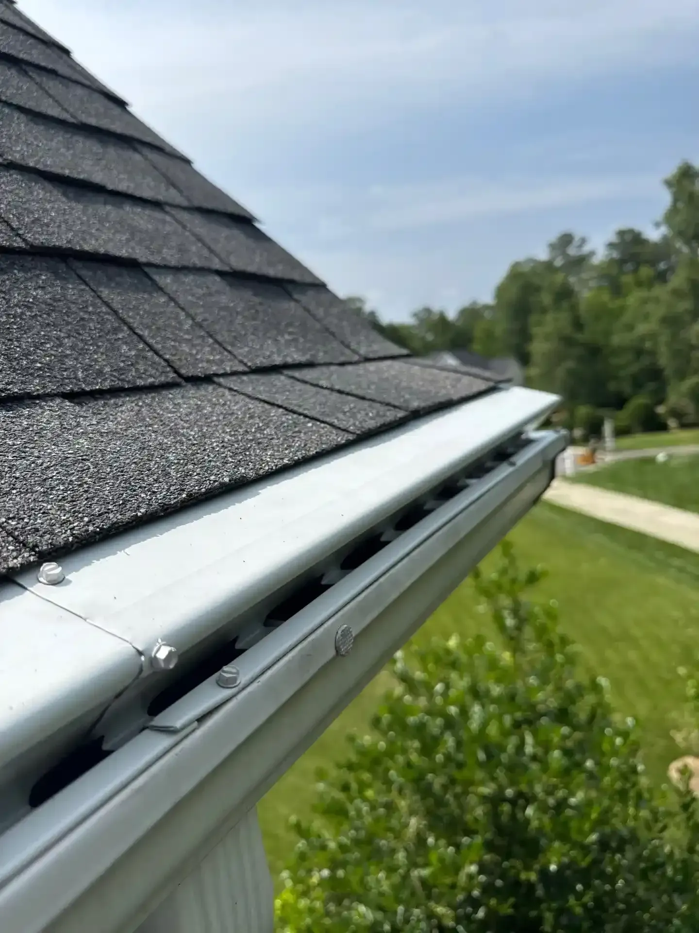 Close-up of gutter guard beneath a dark shingled roof, with trees and a lawn in the background