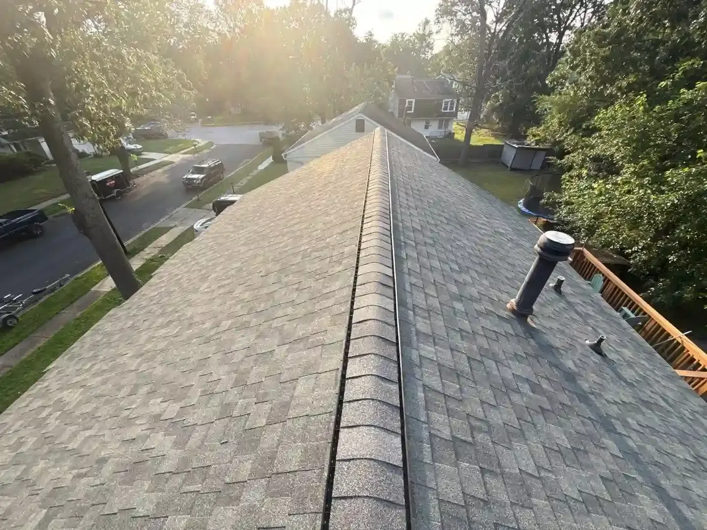 Roofer working on a gray shingle roof ridge beside a gutter, with trees and parked cars below.