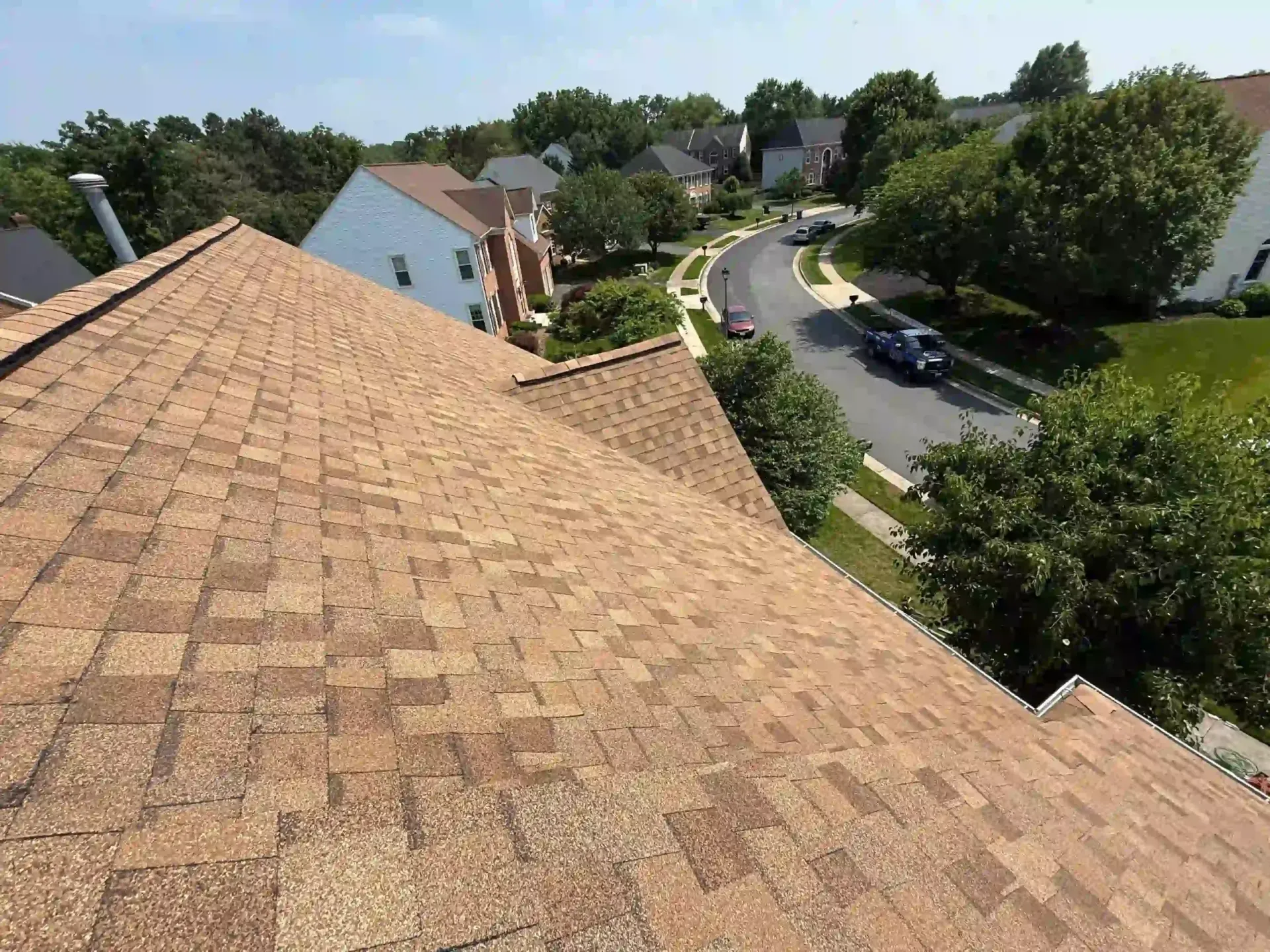View from a shingled roof over a curving suburban street with trees and houses under a bright sky