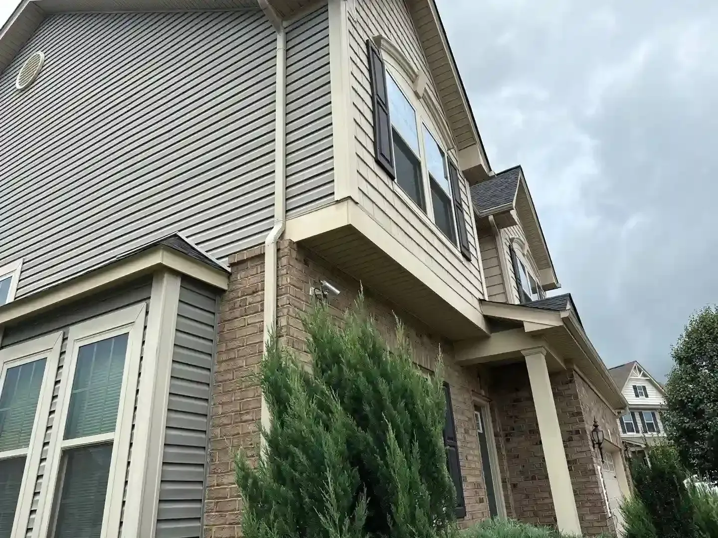 Exterior of a beige townhouse with brick accents, windows, and tall shrubs under a cloudy sky