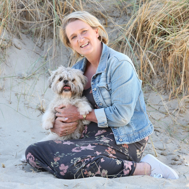 Bibian zit op het strand met het pluizige hondje Binky in haar armen. Ze lachen allebei, in casual kleding. Duinachtergrond
