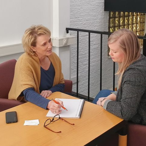 Twee vrouwen zitten aan een tafel. Een van hen wijst naar een papier terwijl zij praat.