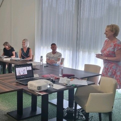 Mensen in een vergadering, een vrouw die een presentatie geeft. Tafels, een laptop met projector, waterflessen. 
