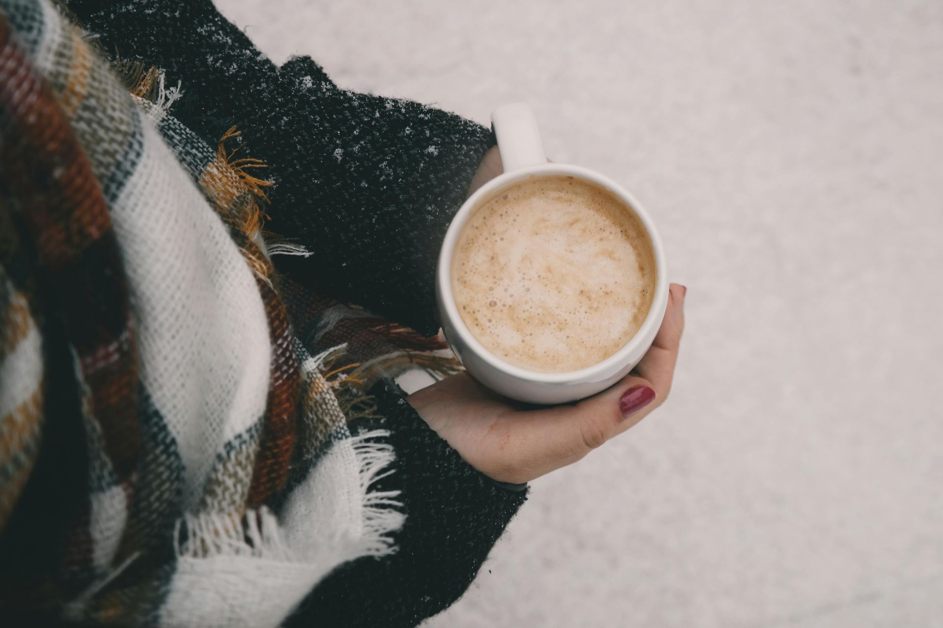 Hand met een mok cappuccino, gewikkeld in een geruite sjaal, tegen een besneeuwde achtergrond.