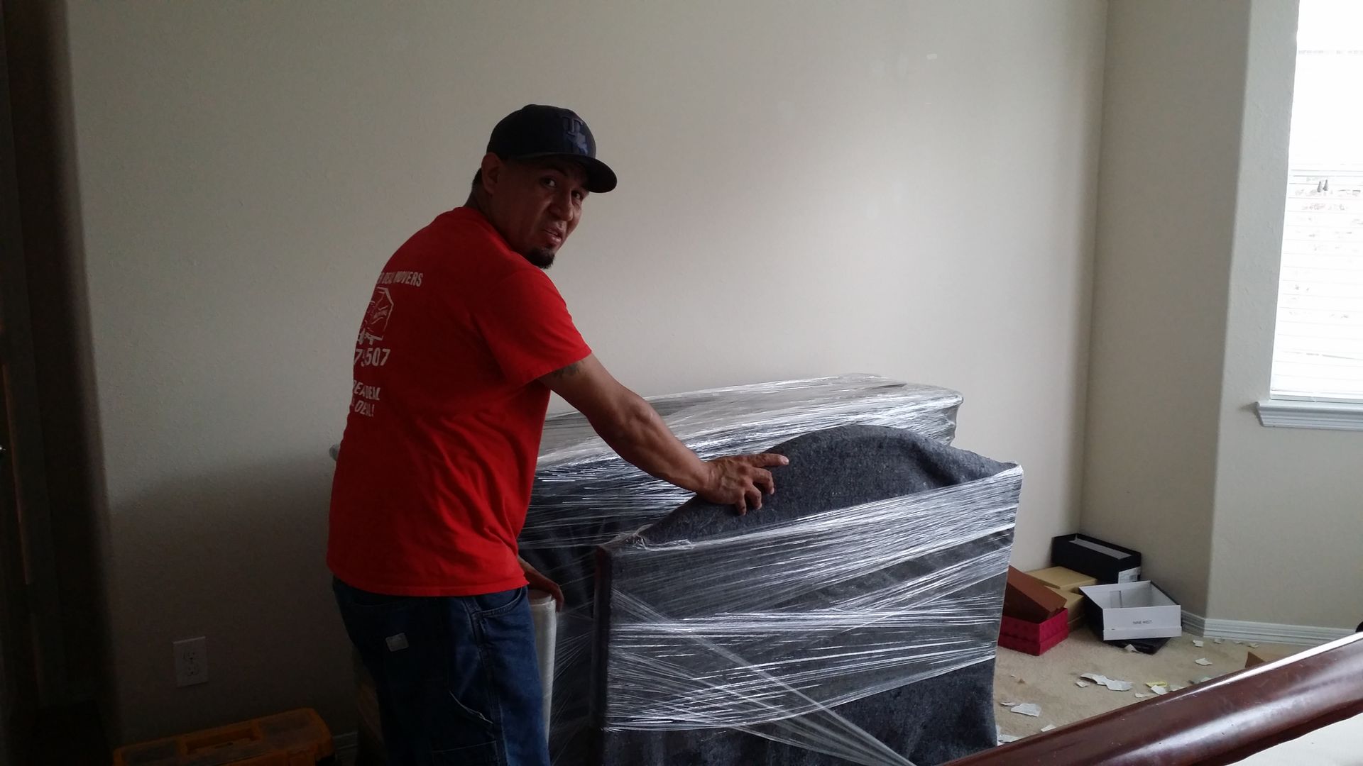 Person in a red shirt unwrapping a large item wrapped in plastic in a room with a window