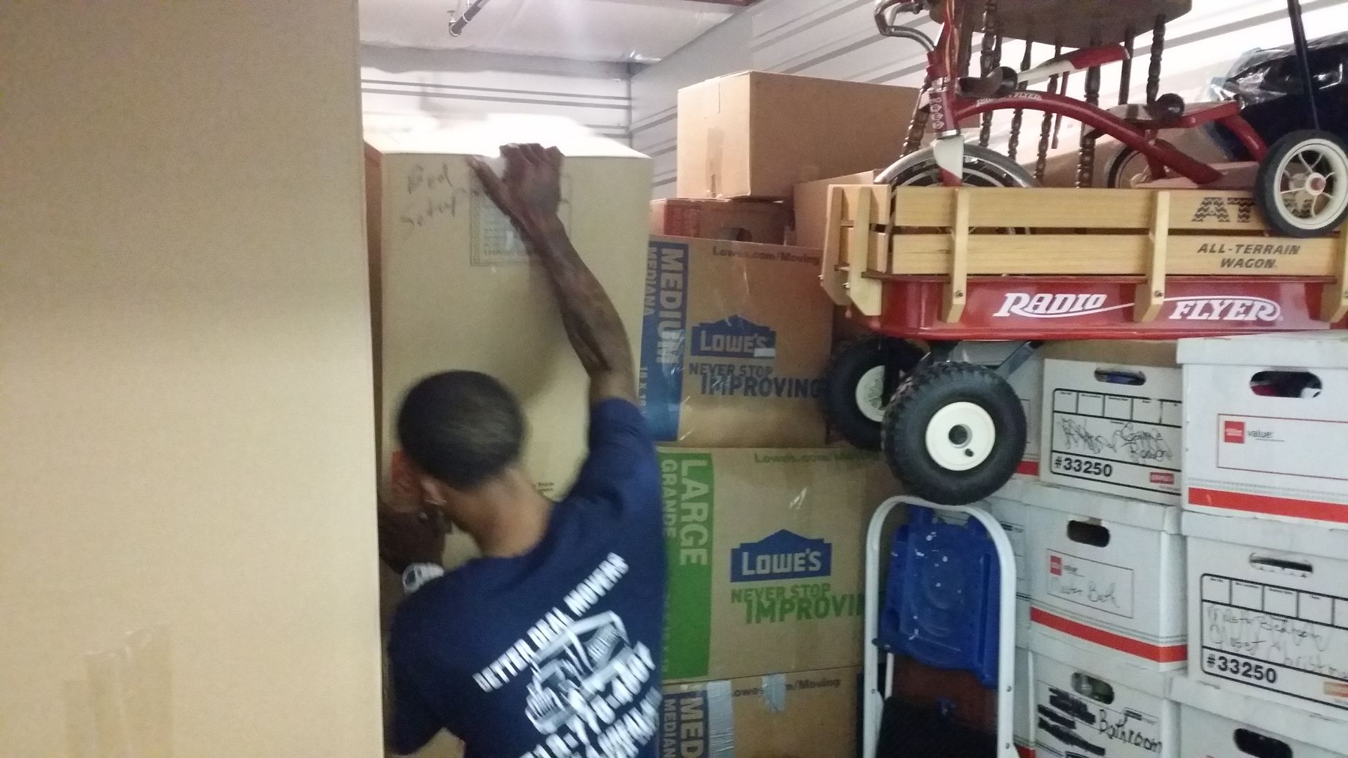 Person lifting cardboard boxes in a packed storage room with stacked boxes and a red wagon.