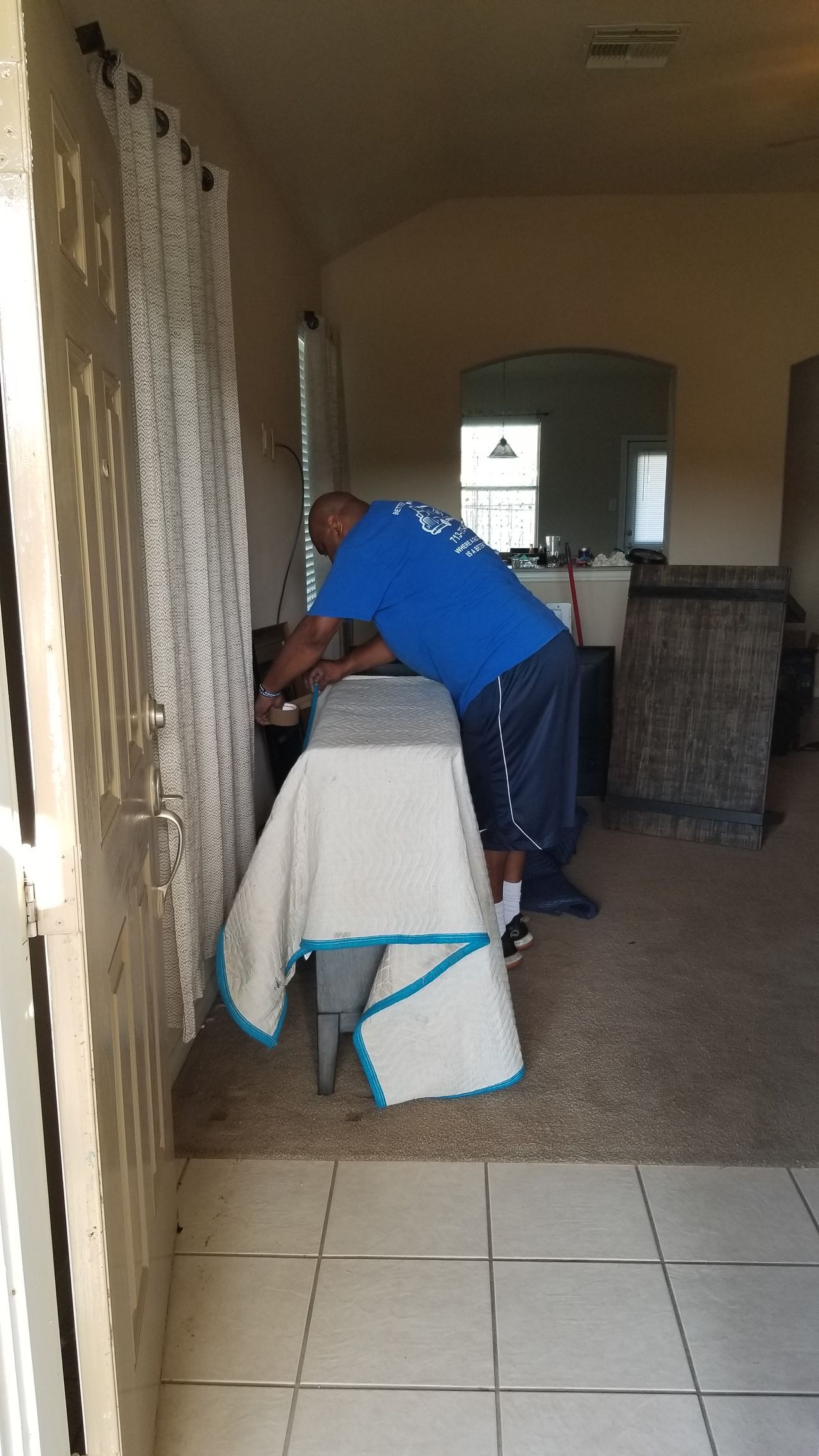 Person in blue shirt leaning over a covered chair by a window in a carpeted room.
