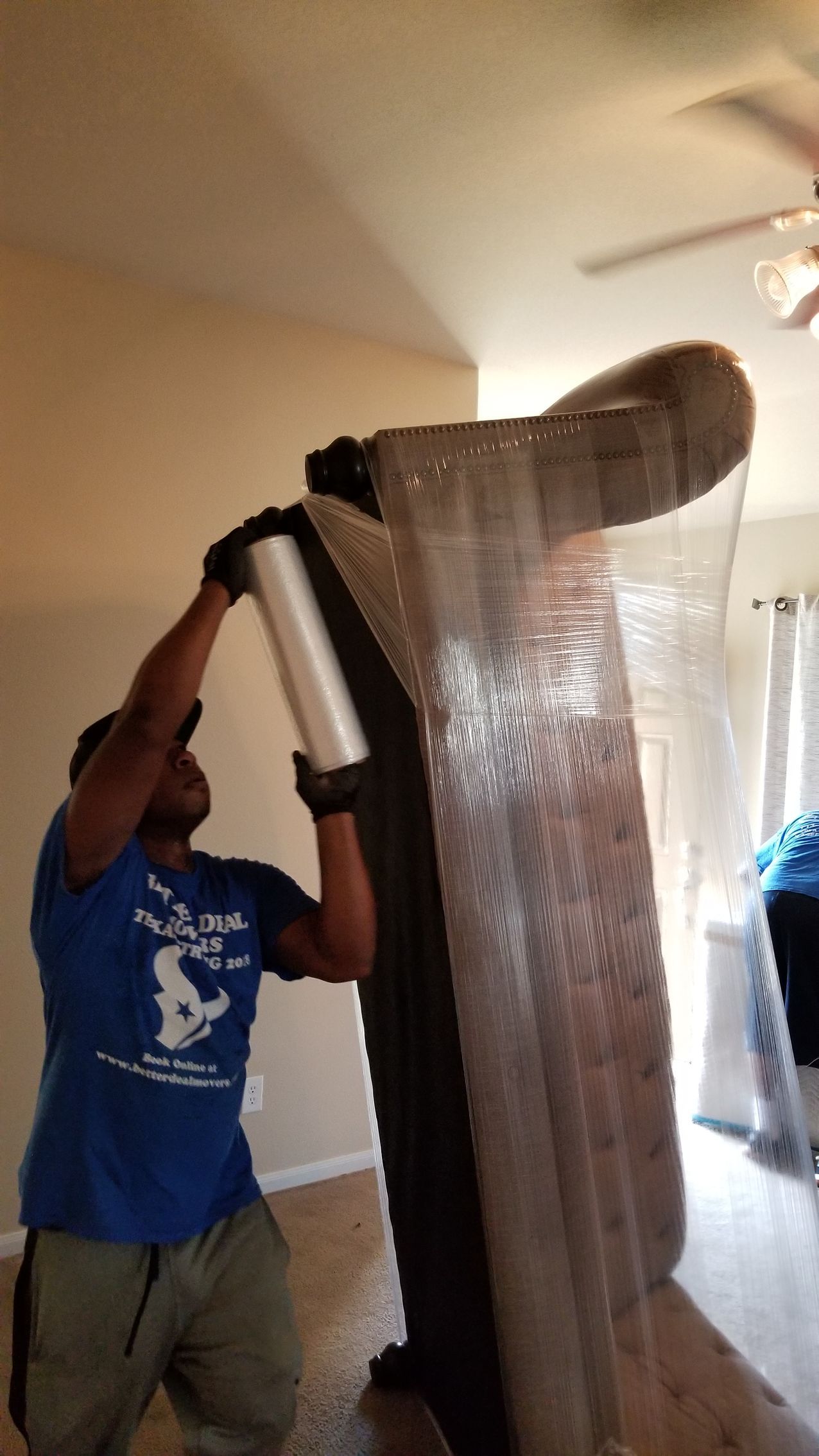 Person wrapping a large mattress in plastic inside a room, with a ceiling fan overhead