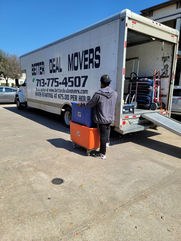 Person carrying an orange suitcase beside a white moving truck on a sunny street
