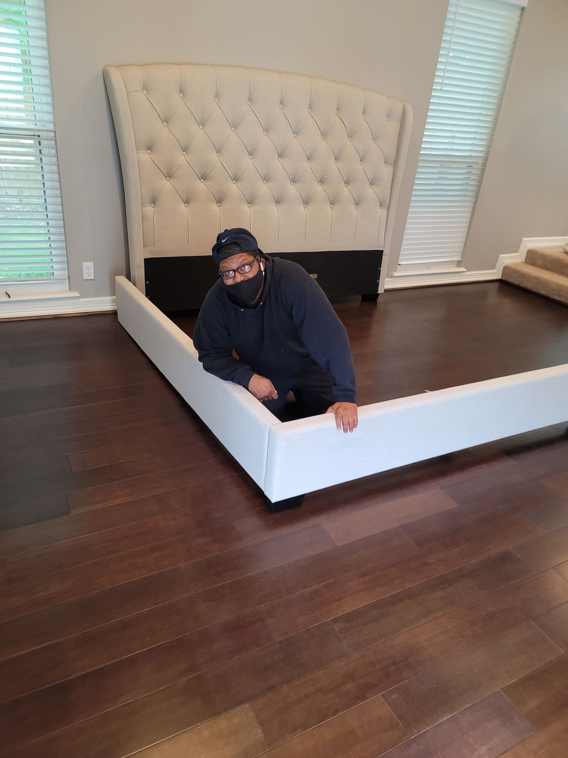 Person kneeling on a white bed frame in a room with dark wood floors and a tufted headboard.