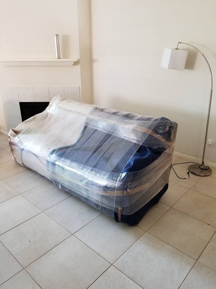 A bed wrapped in plastic in a sparse room with a white floor lamp and tiled floor.