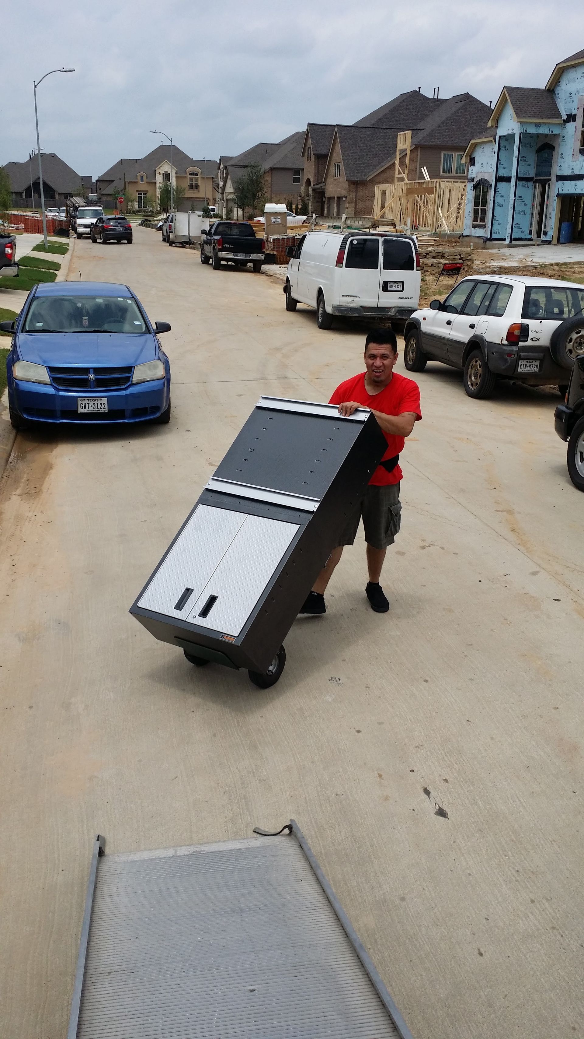 Person pushing a handcart with a panel on a muddy street lined with parked cars and houses