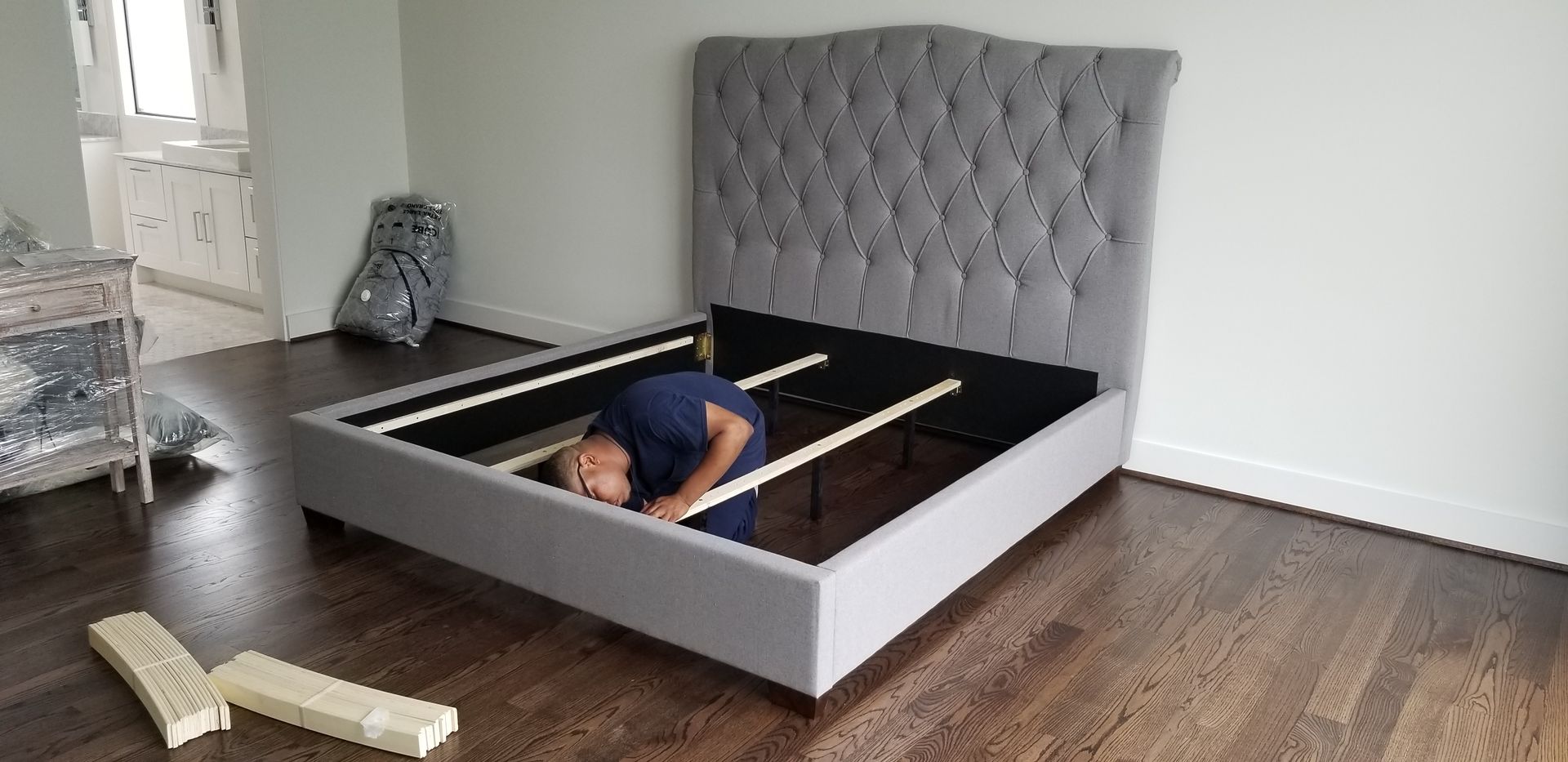 Person assembling a gray upholstered bed frame in a bedroom with wood floors and a white wall