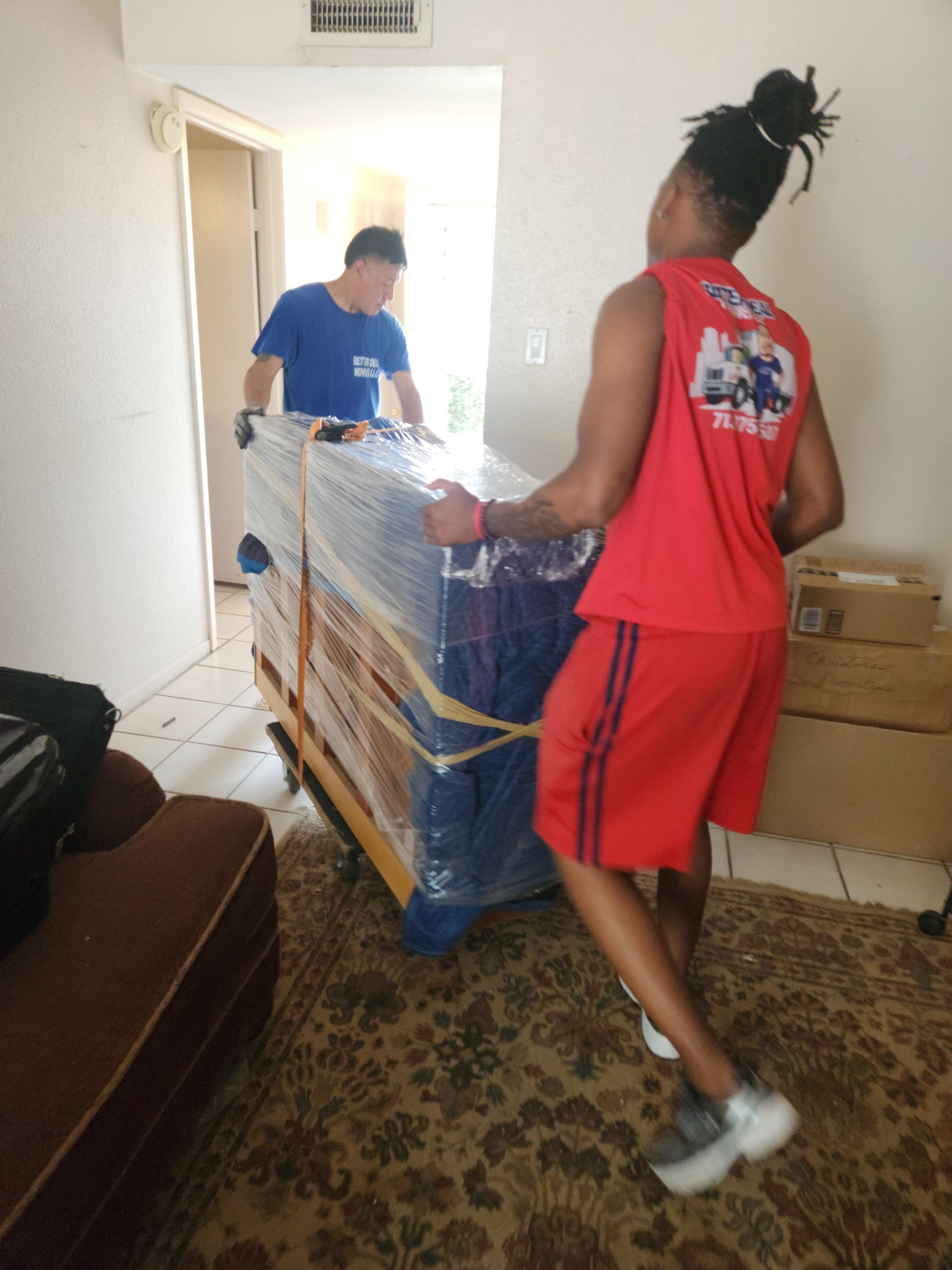 Two people move a wrapped appliance through a doorway in a small room