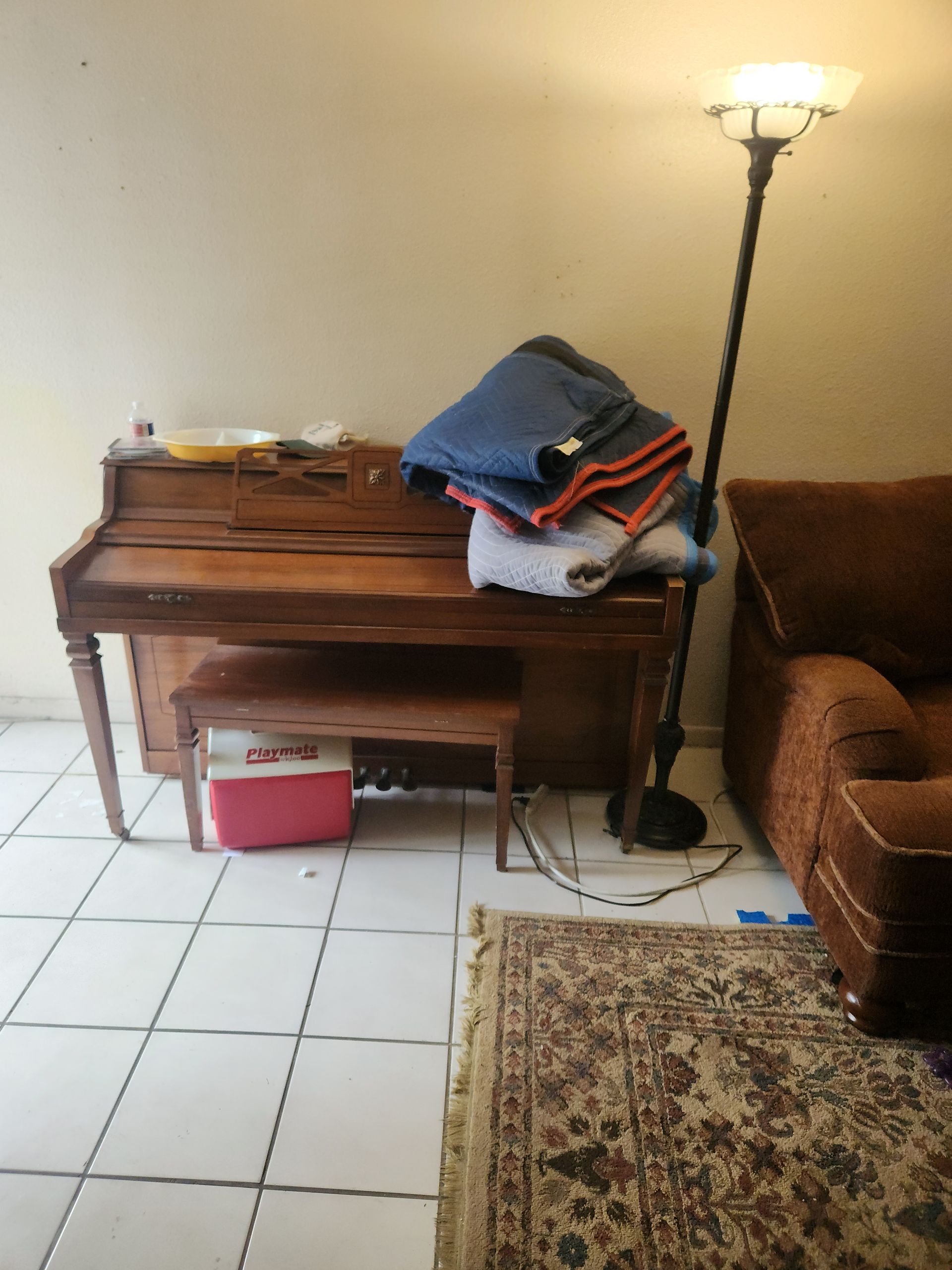 Living room corner with a wooden piano, stacked clothes, floor lamp, armchair, and tiled floor.