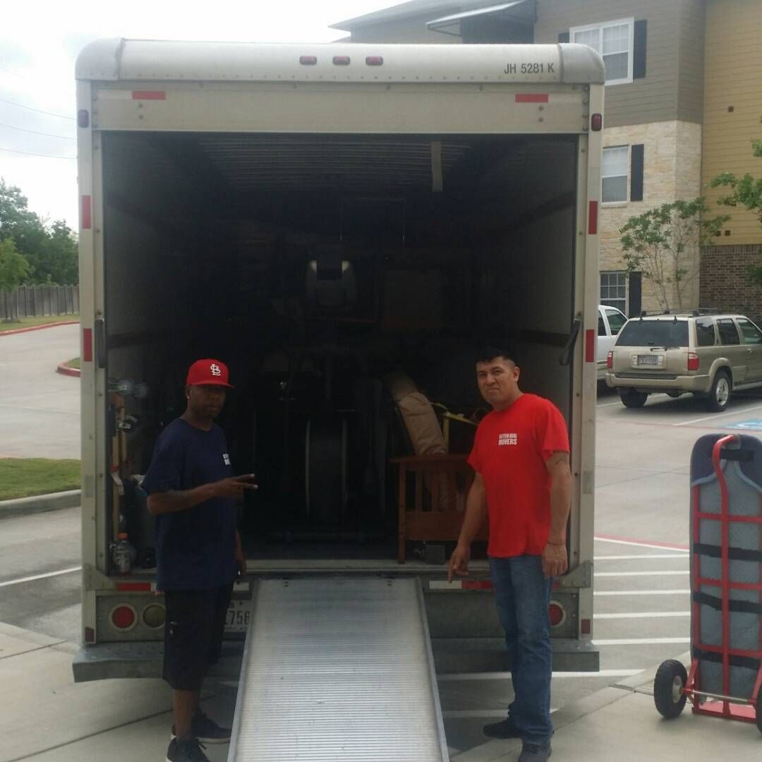 Two men stand by an open moving truck with a ramp outside a building.