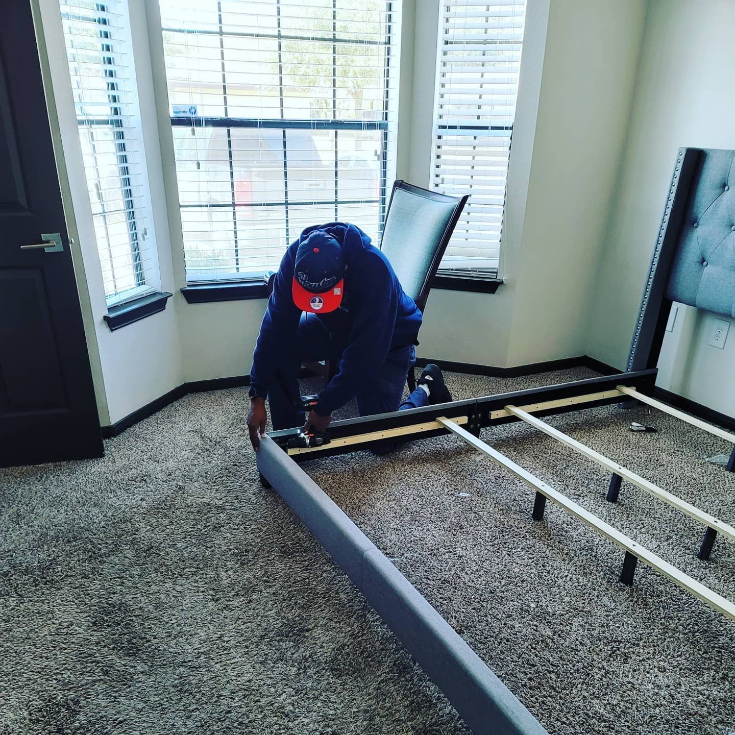 Person assembling a gray bed frame in a carpeted room with windows and shutters
