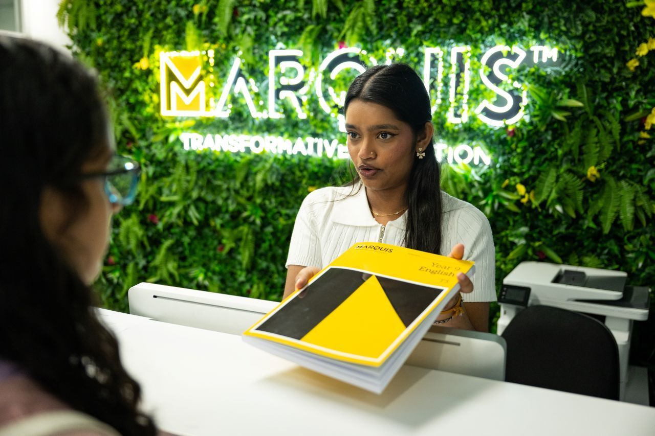 Two women at a reception desk with a yellow-black brochure, in front of a green “MARCUS” wall sign.